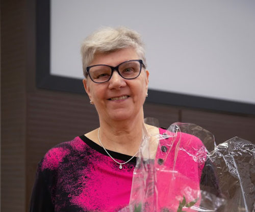 Nainen, harmaat lyhyet hiukset ja silmälasit, hymyilee kukkakimput sylissään.